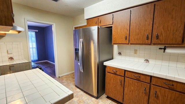 kitchen with backsplash, a textured ceiling, tile counters, exhaust hood, and stainless steel refrigerator with ice dispenser