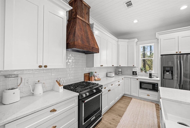 kitchen with white cabinets, light wood-type flooring, custom exhaust hood, appliances with stainless steel finishes, and light stone counters