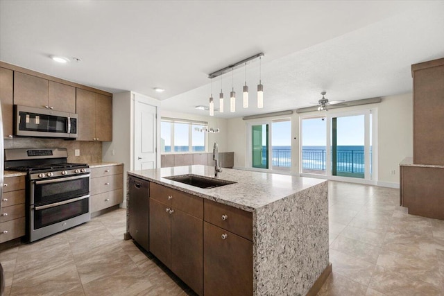 kitchen with stainless steel appliances, a water view, a kitchen island with sink, a sink, and light stone countertops