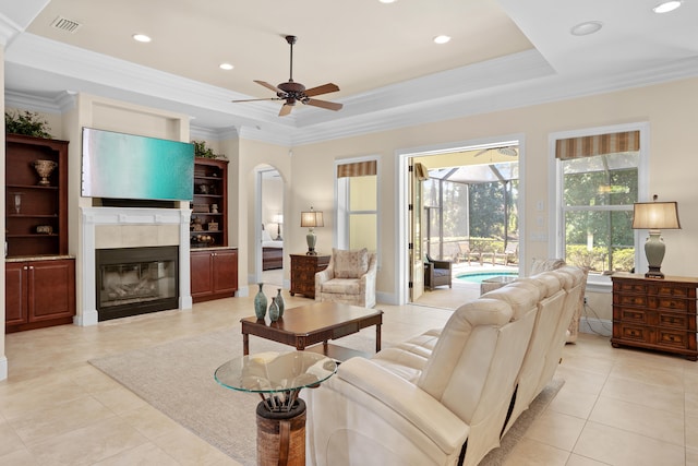 living room featuring a raised ceiling, a tile fireplace, ceiling fan, crown molding, and light tile patterned floors