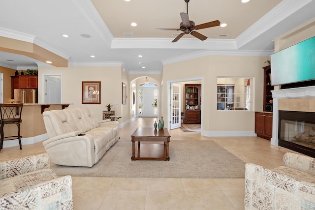 tiled living room with a tiled fireplace, ornamental molding, a tray ceiling, and ceiling fan