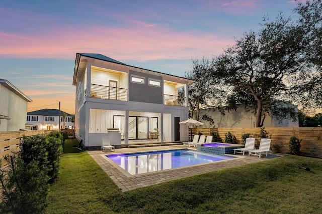 back house at dusk featuring a lawn, a balcony, a patio, and a pool with hot tub