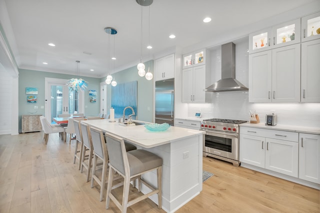 kitchen featuring wall chimney range hood, an island with sink, hanging light fixtures, high end appliances, and white cabinetry