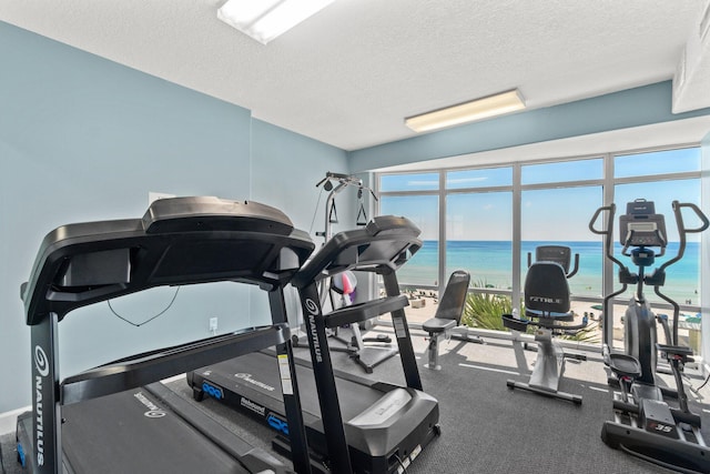 gym featuring a textured ceiling, a water view, and a beach view