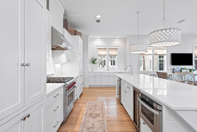 kitchen featuring light hardwood / wood-style flooring, wall chimney range hood, white cabinetry, appliances with stainless steel finishes, and decorative light fixtures