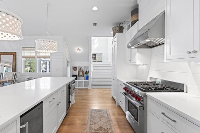 kitchen featuring luxury stove, wall chimney range hood, white cabinets, and hanging light fixtures