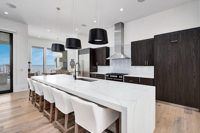 kitchen with a kitchen island with sink, a water view, wall chimney range hood, light wood-type flooring, and decorative light fixtures