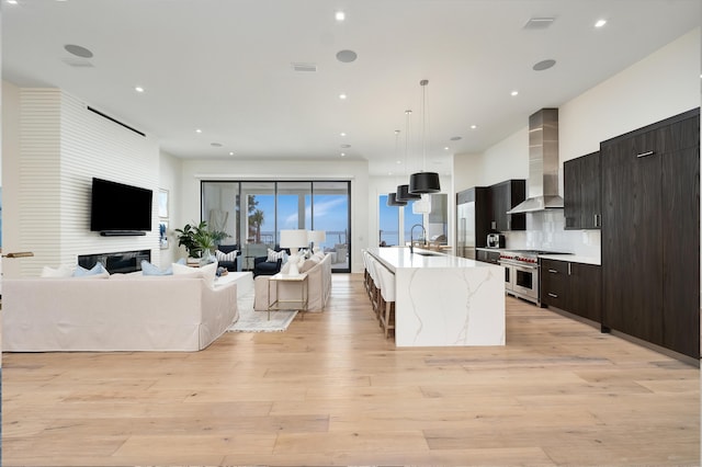 kitchen featuring wall chimney exhaust hood, decorative light fixtures, light wood-type flooring, a large island with sink, and double oven range