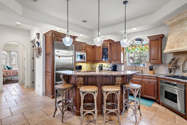 kitchen featuring a kitchen island, backsplash, light stone countertops, built in appliances, and pendant lighting