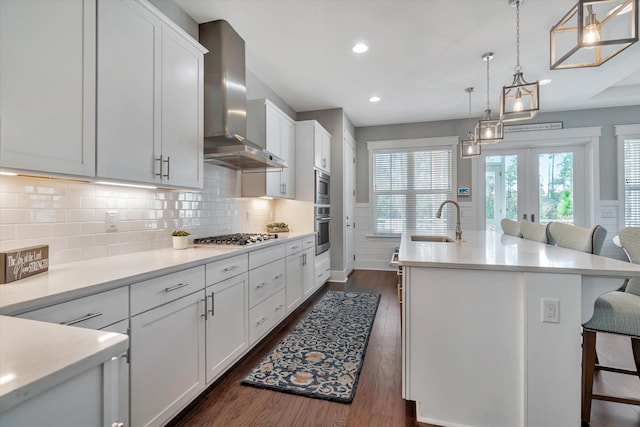 kitchen with wall chimney range hood, appliances with stainless steel finishes, a kitchen island with sink, white cabinetry, and a kitchen breakfast bar