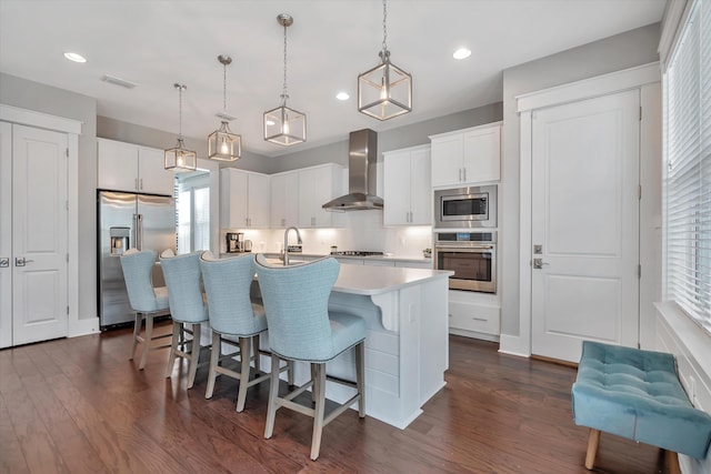 kitchen with a center island with sink, hanging light fixtures, appliances with stainless steel finishes, wall chimney range hood, and white cabinets