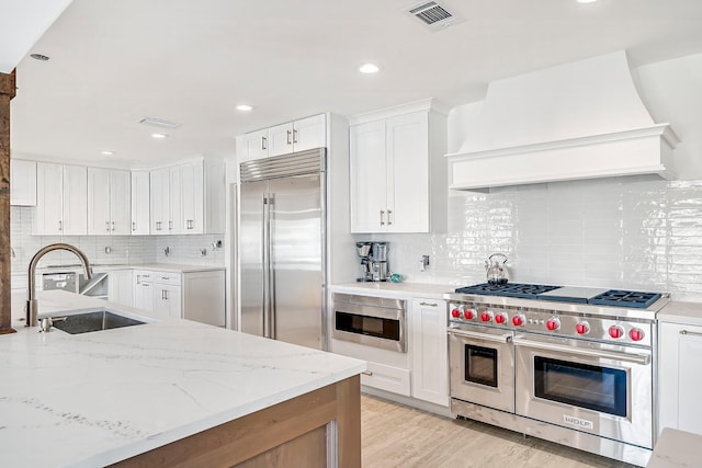 kitchen with custom range hood, high end appliances, sink, white cabinetry, and light stone counters