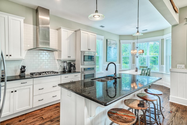 kitchen featuring wall chimney range hood, a center island with sink, appliances with stainless steel finishes, dark wood-type flooring, and pendant lighting