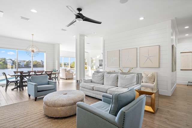 living room featuring a water view, ceiling fan with notable chandelier, decorative columns, wooden walls, and light wood-type flooring