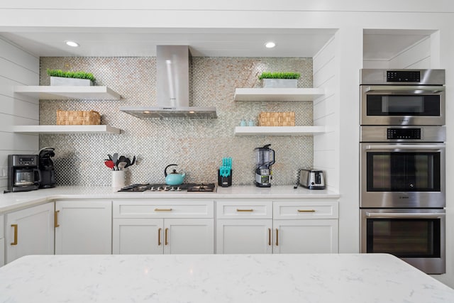 kitchen featuring light stone counters, appliances with stainless steel finishes, decorative backsplash, white cabinets, and wall chimney exhaust hood