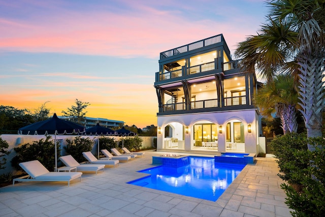 back house at dusk featuring a swimming pool with hot tub, a patio, and a balcony