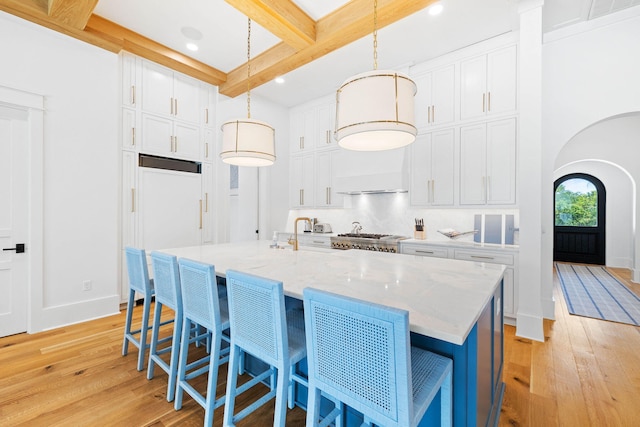 kitchen with light wood-type flooring, decorative light fixtures, beam ceiling, white cabinets, and a kitchen island with sink