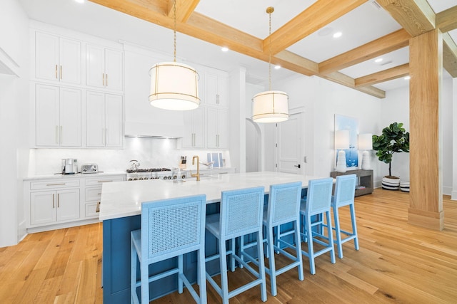 kitchen with white cabinetry, light wood-type flooring, beam ceiling, hanging light fixtures, and a kitchen island with sink