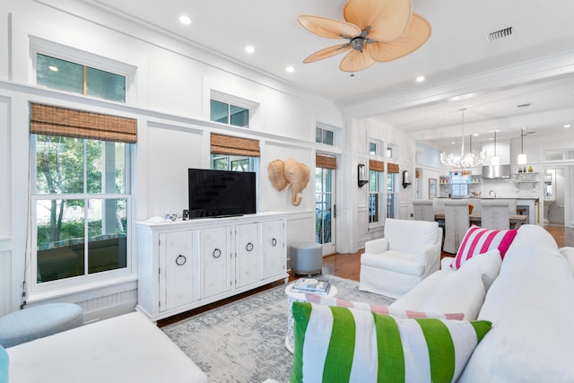 living room with light wood-type flooring, beamed ceiling, ceiling fan with notable chandelier, and crown molding