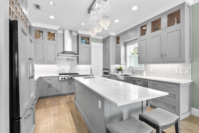 kitchen featuring ornamental molding, a center island with sink, stainless steel appliances, wall chimney range hood, and decorative light fixtures