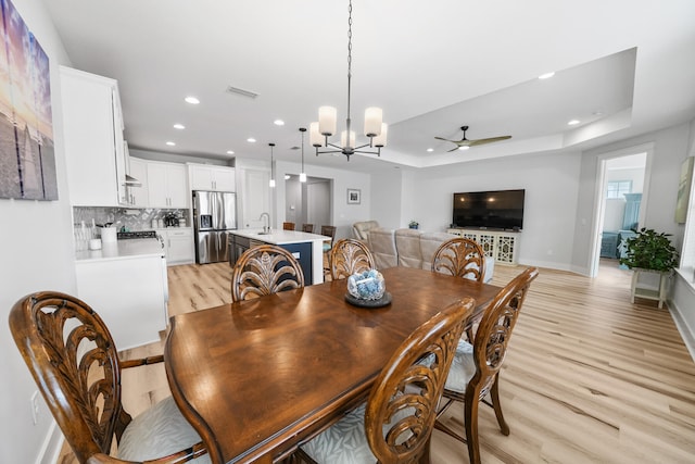 dining space featuring ceiling fan with notable chandelier, light hardwood / wood-style flooring, sink, and a tray ceiling