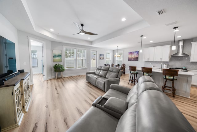 living room with light hardwood / wood-style floors, ceiling fan with notable chandelier, and a raised ceiling