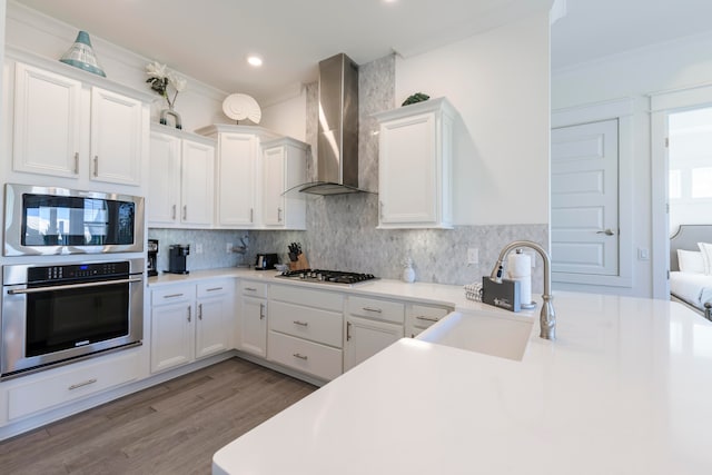 kitchen with ornamental molding, light wood-type flooring, wall chimney range hood, appliances with stainless steel finishes, and white cabinets