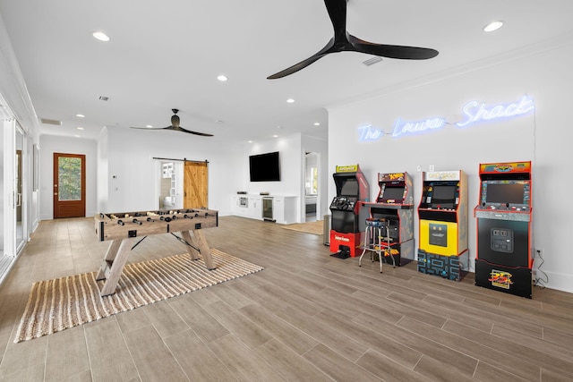 rec room with light wood-type flooring, a barn door, ceiling fan, and ornamental molding