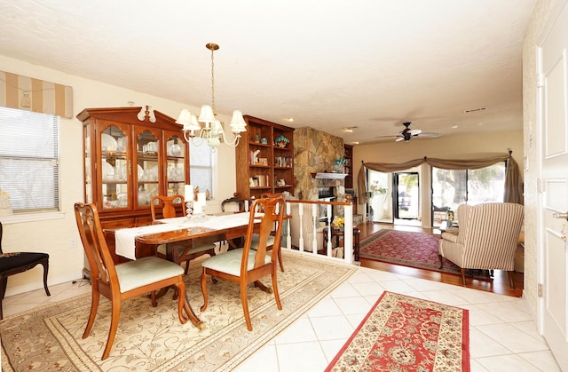 dining space with light tile patterned floors, a stone fireplace, and ceiling fan with notable chandelier