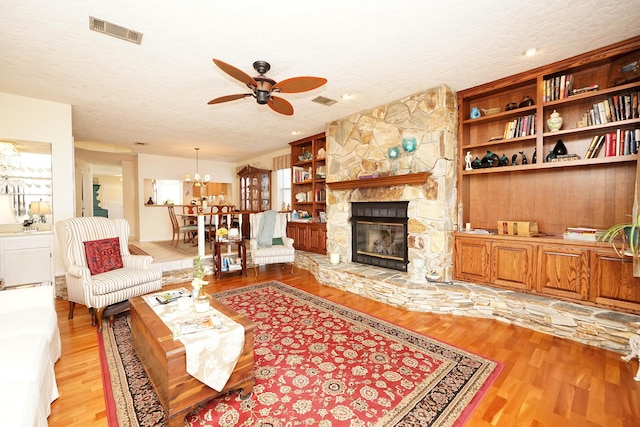 living room featuring a stone fireplace, ceiling fan with notable chandelier, a textured ceiling, and light wood-type flooring