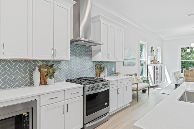 kitchen with white cabinetry, ornamental molding, wall chimney range hood, plenty of natural light, and appliances with stainless steel finishes