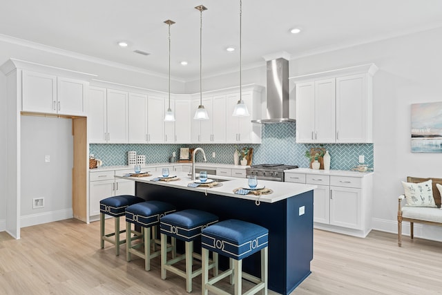 kitchen with decorative light fixtures, white cabinets, high end stove, wall chimney exhaust hood, and a kitchen island with sink