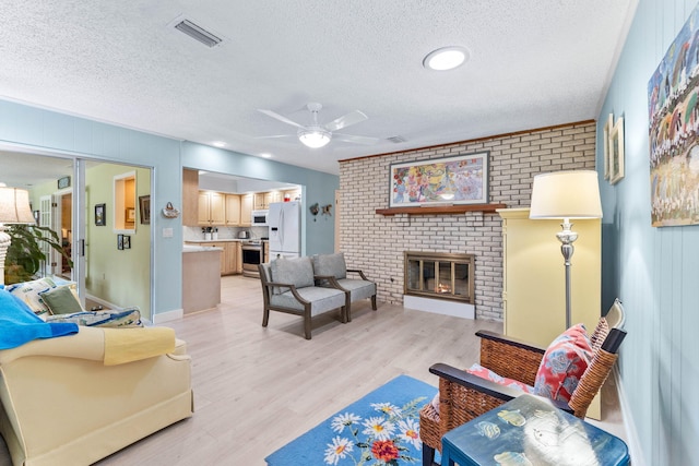 living room with a brick fireplace, a textured ceiling, light hardwood / wood-style flooring, and ceiling fan