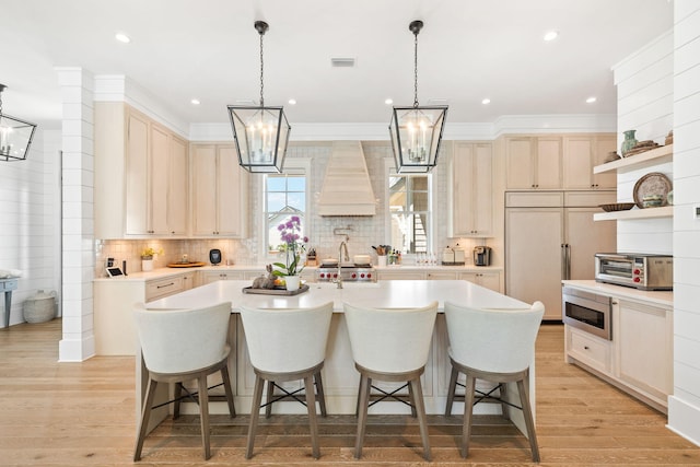 kitchen featuring light wood-type flooring, ornamental molding, custom exhaust hood, a large island with sink, and built in appliances