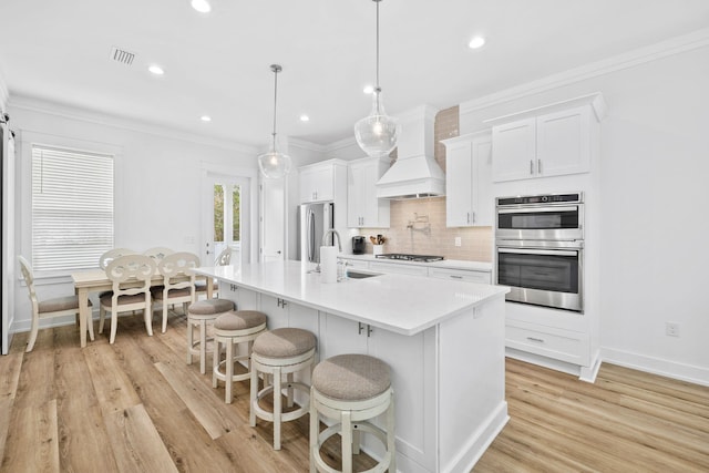 kitchen featuring stainless steel appliances, pendant lighting, an island with sink, white cabinets, and custom exhaust hood