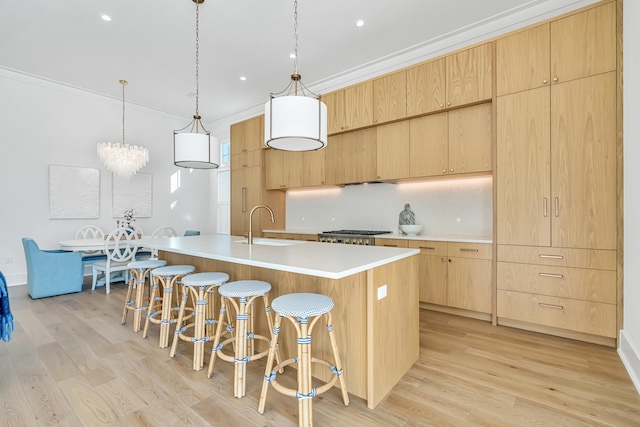 kitchen featuring light wood-type flooring, crown molding, light brown cabinets, hanging light fixtures, and an island with sink