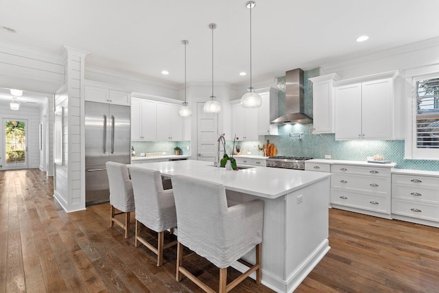 kitchen featuring wall chimney exhaust hood, a breakfast bar, stainless steel appliances, a center island with sink, and white cabinetry