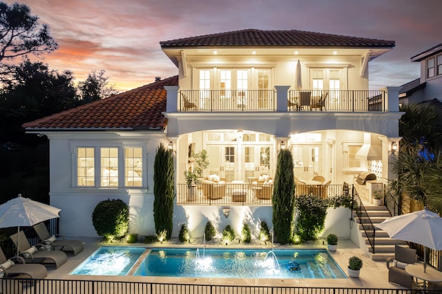 back house at dusk with french doors, a balcony, pool water feature, a swimming pool with hot tub, and a patio area