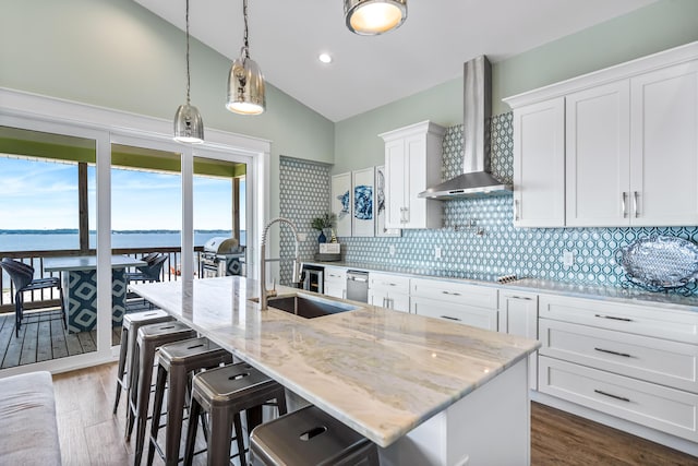 kitchen with white cabinetry, a kitchen island with sink, light stone countertops, a water view, and wall chimney range hood