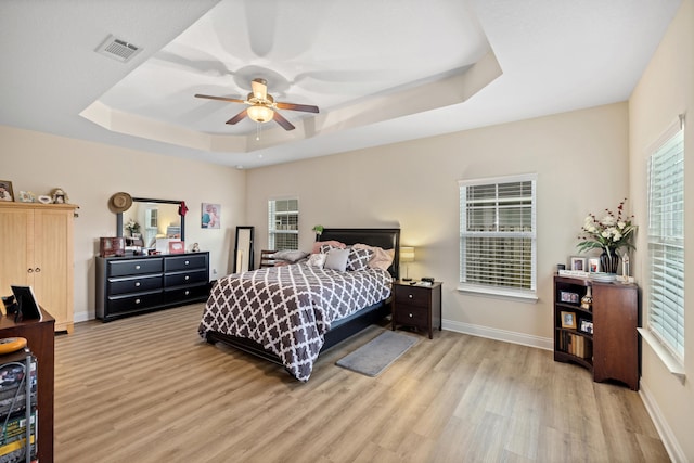 bedroom with ceiling fan, a raised ceiling, light wood-type flooring, and multiple windows