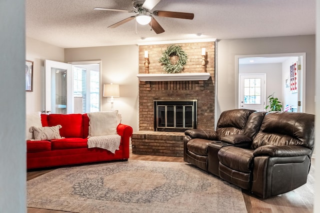 living room with wood-type flooring, a textured ceiling, a brick fireplace, and ceiling fan