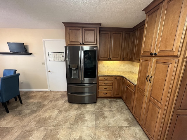 kitchen featuring decorative backsplash, brown cabinets, light stone countertops, a textured ceiling, and stainless steel refrigerator with ice dispenser