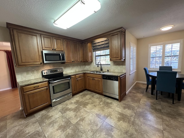 kitchen with light stone counters, stainless steel appliances, a sink, backsplash, and brown cabinets