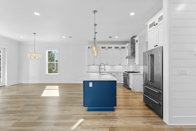 kitchen featuring wall chimney range hood, a center island with sink, high quality appliances, white cabinetry, and hanging light fixtures