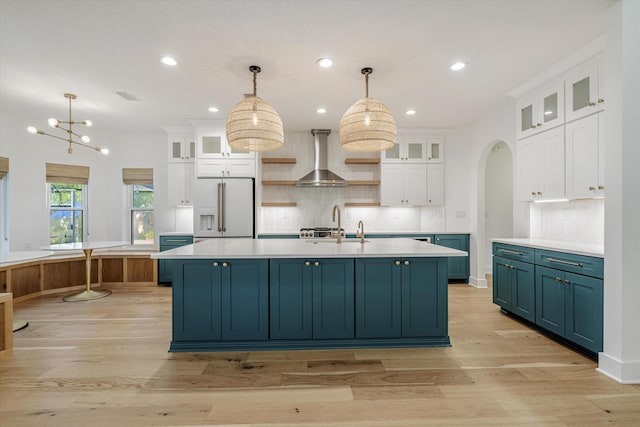 kitchen featuring white cabinetry, wall chimney range hood, white refrigerator with ice dispenser, an island with sink, and decorative light fixtures