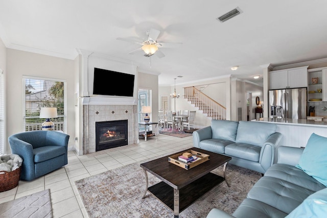 living room with ceiling fan with notable chandelier, light tile patterned floors, crown molding, and a tiled fireplace