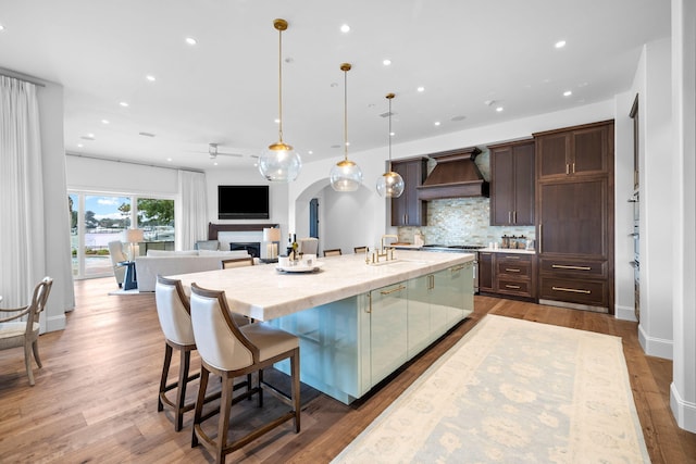 kitchen featuring hardwood / wood-style floors, premium range hood, a center island with sink, decorative light fixtures, and dark brown cabinets