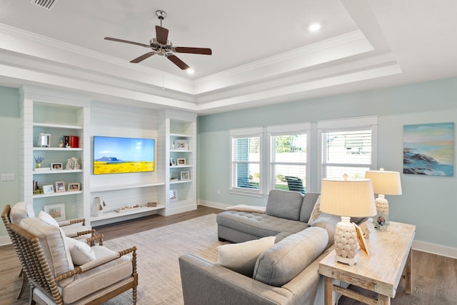 living room featuring built in shelves, a tray ceiling, ceiling fan, crown molding, and wood-type flooring