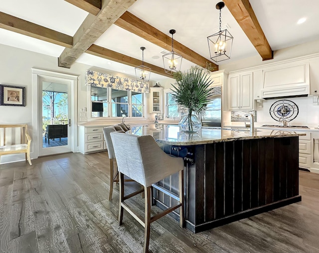 kitchen featuring backsplash, light stone counters, a kitchen island with sink, decorative light fixtures, and white cabinetry