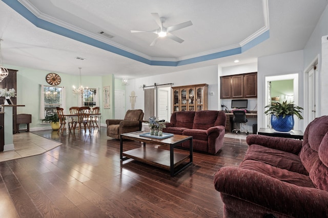 living room featuring dark hardwood / wood-style flooring, ornamental molding, ceiling fan with notable chandelier, a tray ceiling, and a barn door